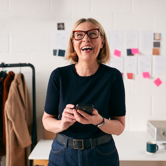 Laughing Mature Businesswoman With Mobile Phone In Front Of Desk In Start Up Fashion Business